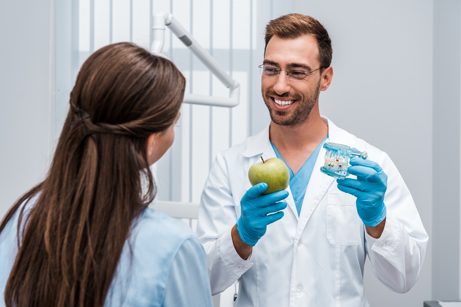 dentist in glasses holding tooth model near patient